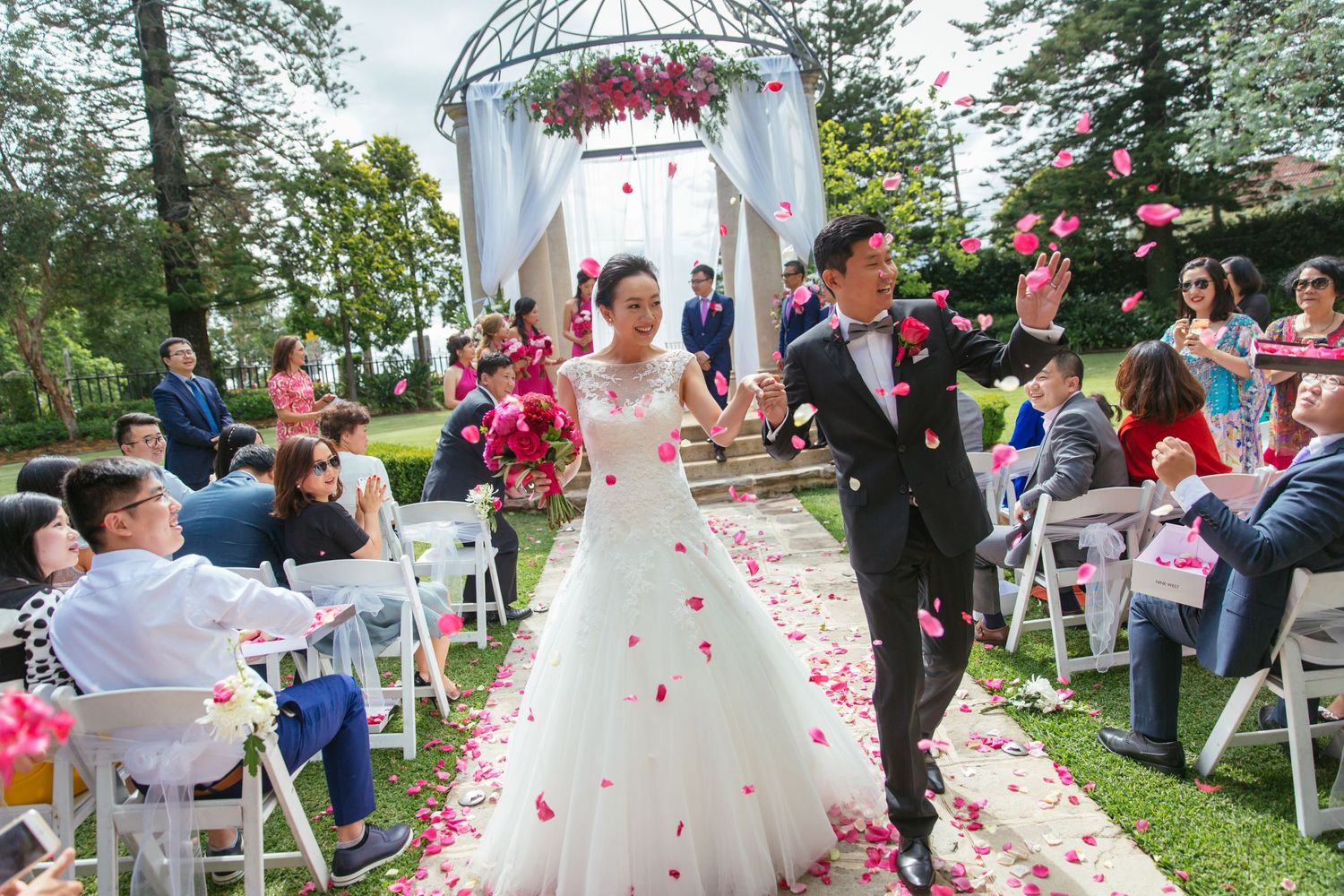 Joyful newlyweds walk down floral arch aisle as guests shower them with pink rose petals outdoors.