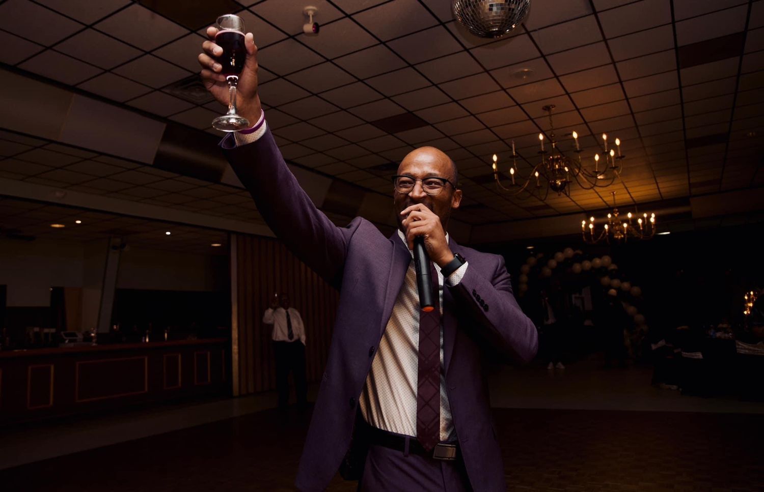 Person in purple suit jacket holding up a glass for a toast while speaking into microphone.