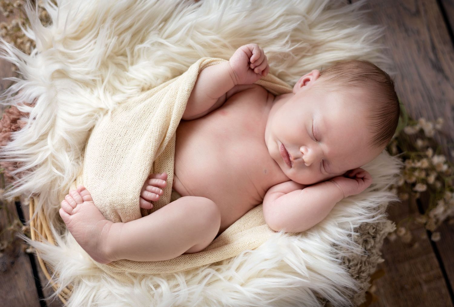 Sleeping newborn baby wrapped in cream blanket lying on white fluffy fur backdrop.