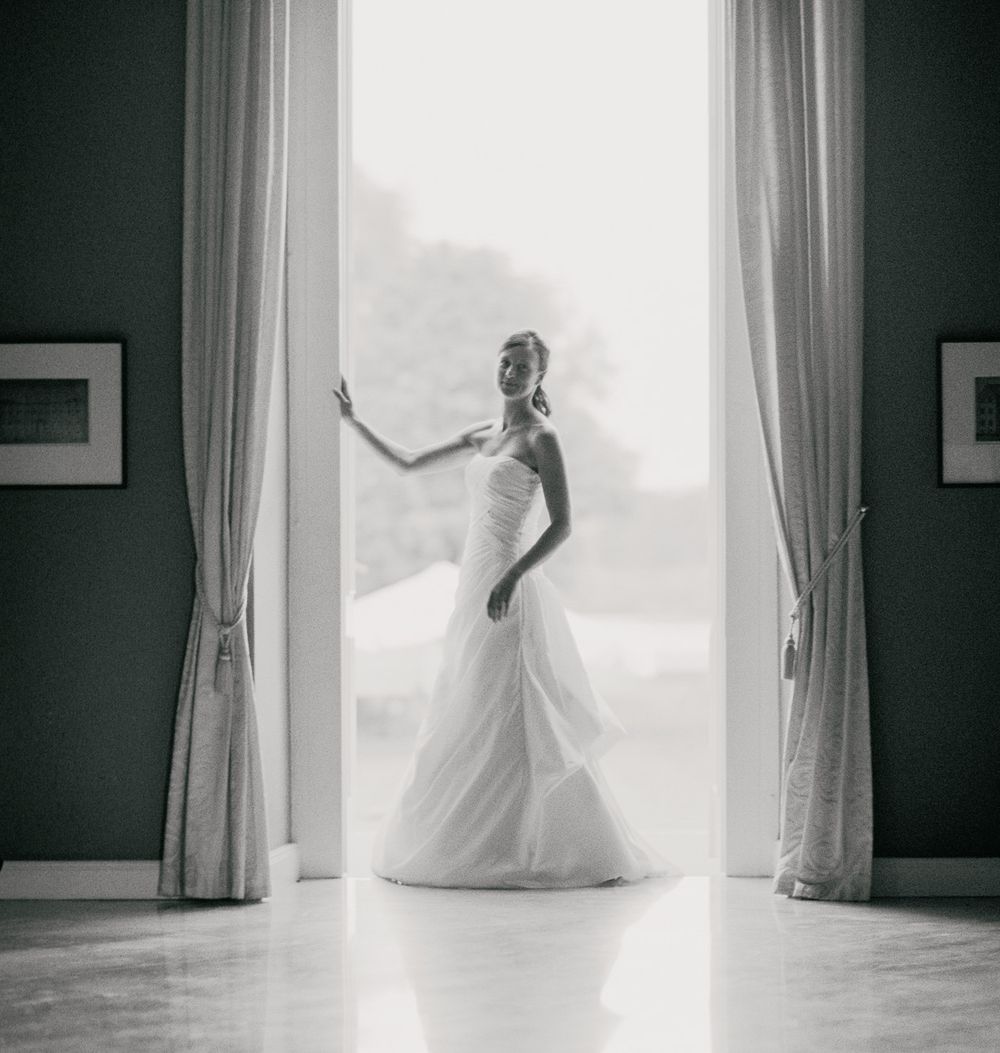 Silhouette of person in wedding dress standing in bright window frame against dark walls.