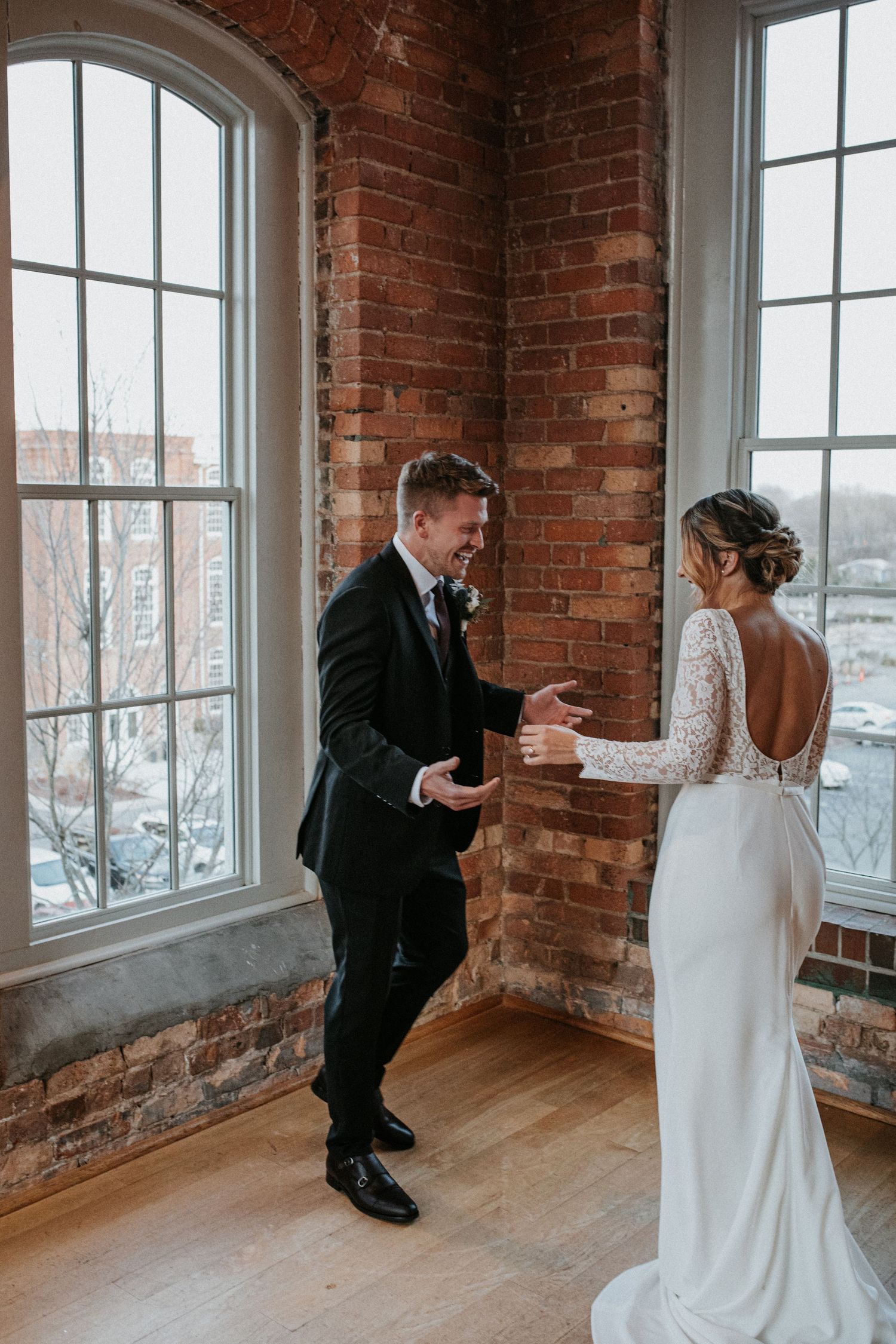A bride in a backless lace gown and groom in black suit share a candid moment by large windows in a rustic brick venue.