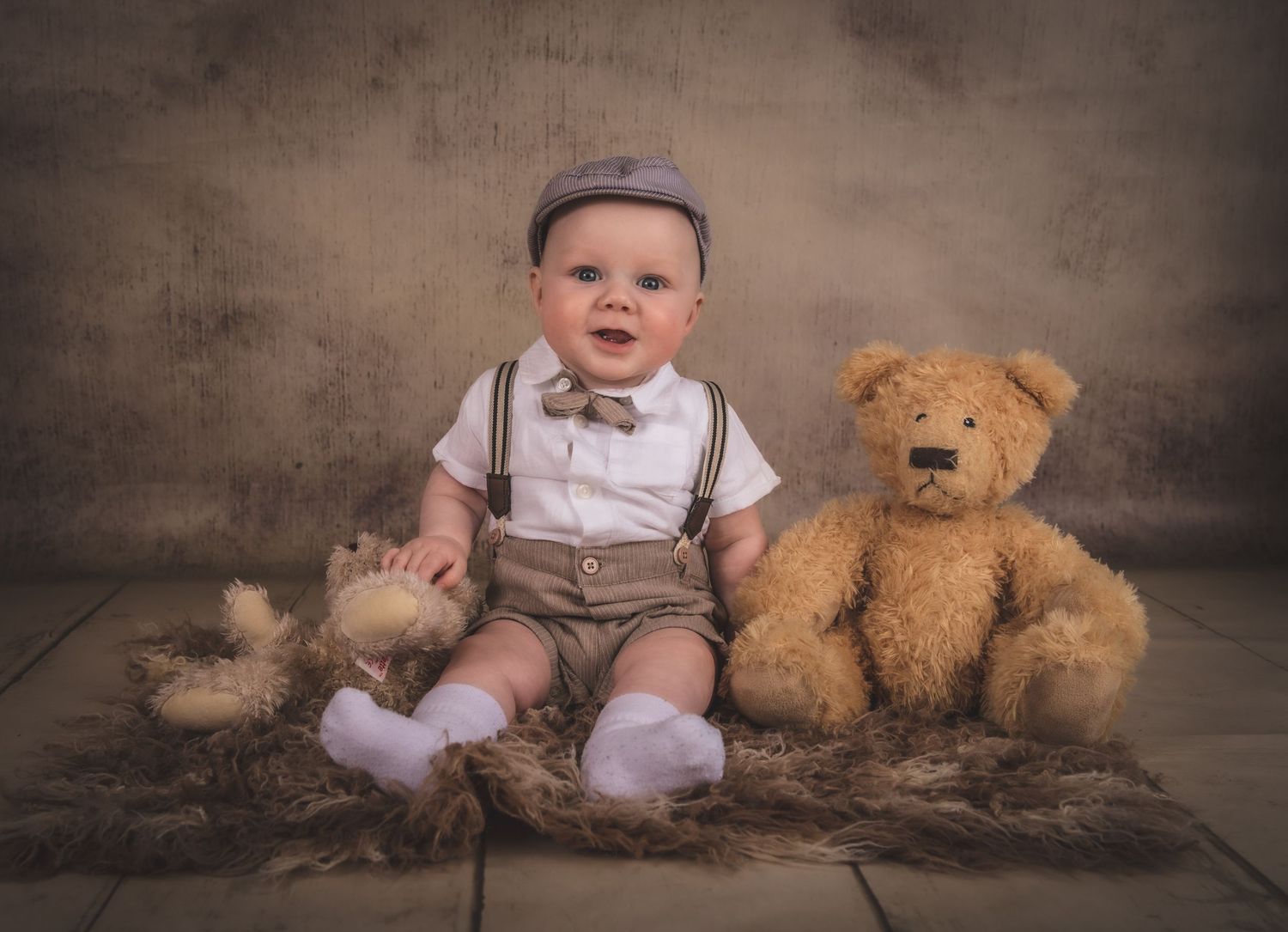 Baby in vintage-style outfit sits with teddy bear against textured backdrop.