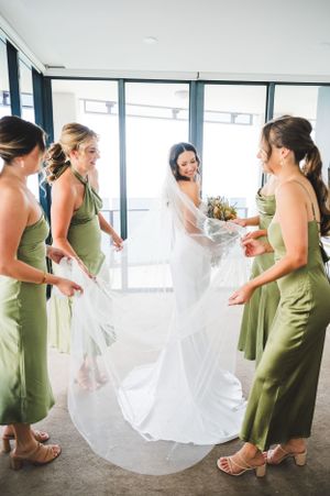 Bridesmaids in sage green dresses gather around a bride in a flowing white gown.