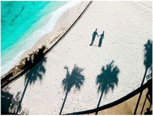 Aerial view of two silhouettes walking on a white sandy beach with palm tree shadows and turquoise water.