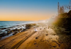 Silhouette on rocky beach shoreline during golden sunset with ocean waves and pine trees.