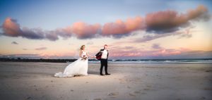 A couple walks along a sandy beach at sunset with pink clouds overhead.