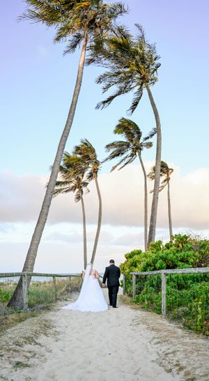 A couple walks down a sandy beach path lined with tall swaying palm trees on their wedding day.