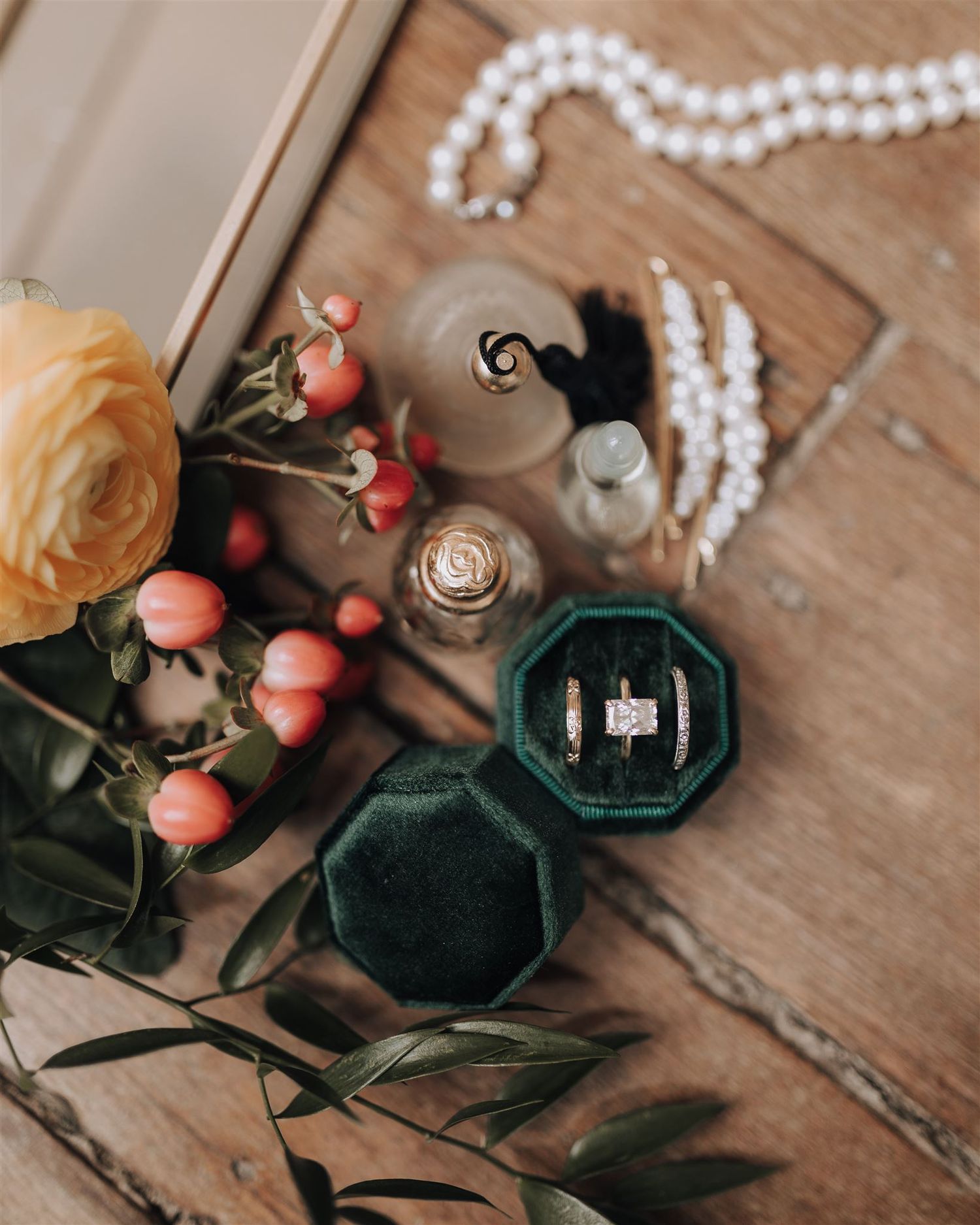 Wedding details flatlay with coral roses, pearl necklace, and emerald ring box on wooden surface.