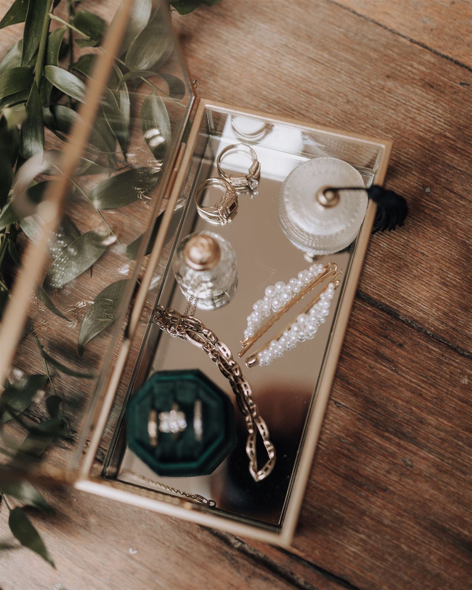 Wedding accessories including jewelry, pearls and perfume bottle arranged on mirrored tray with greenery.