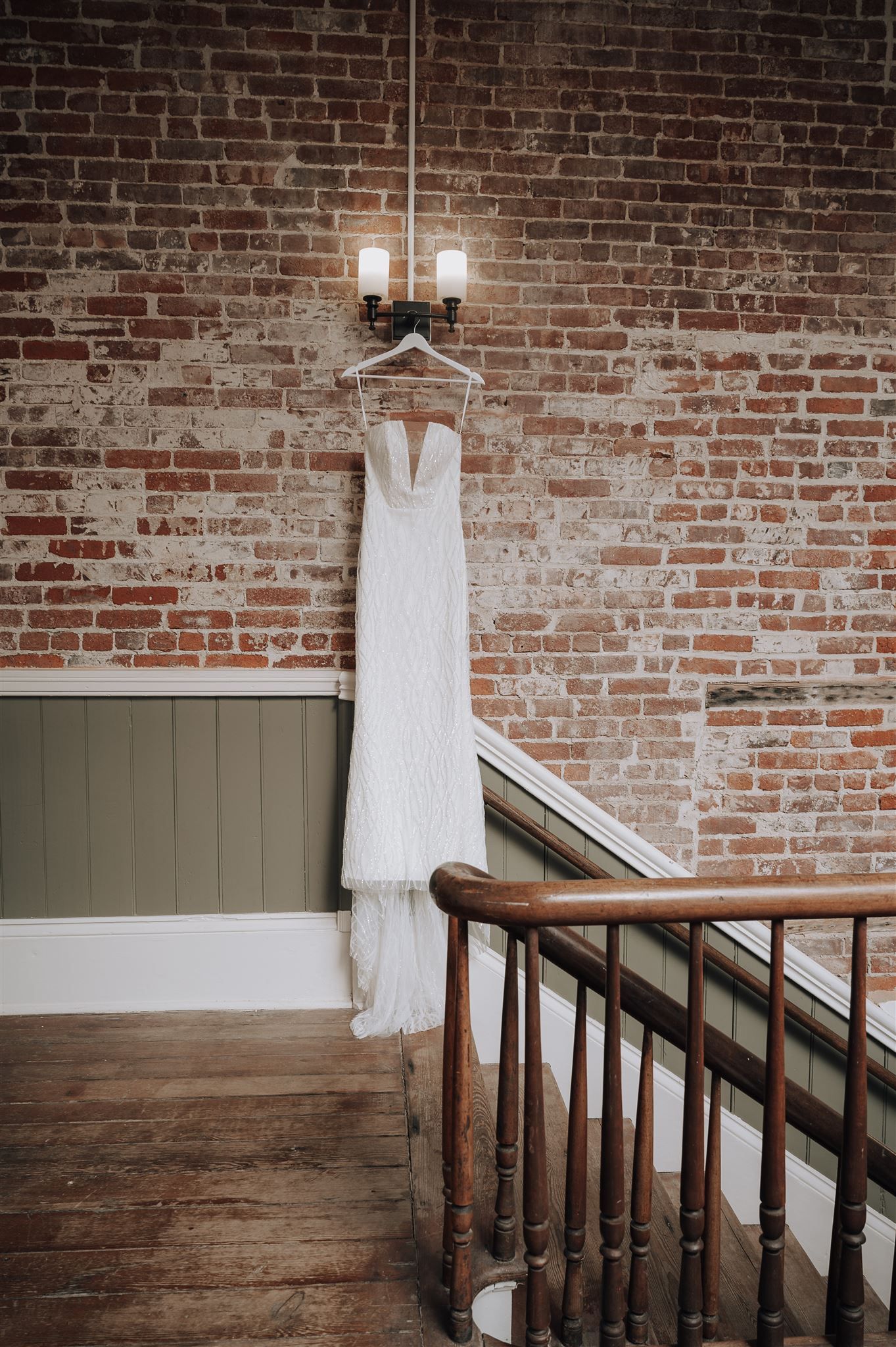 Elegant white wedding gown displayed on hanger against rustic brick wall near wooden staircase.