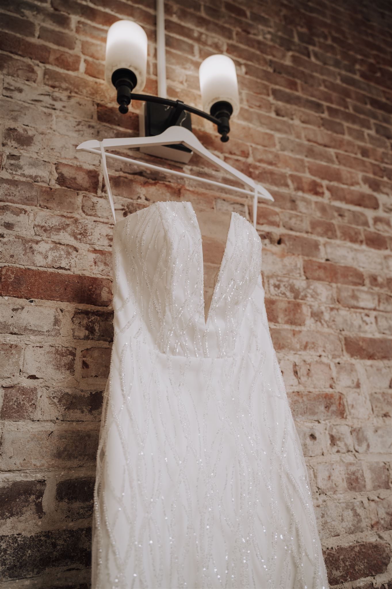 White lace wedding dress hanging on display against exposed brick wall with vintage light fixtures.