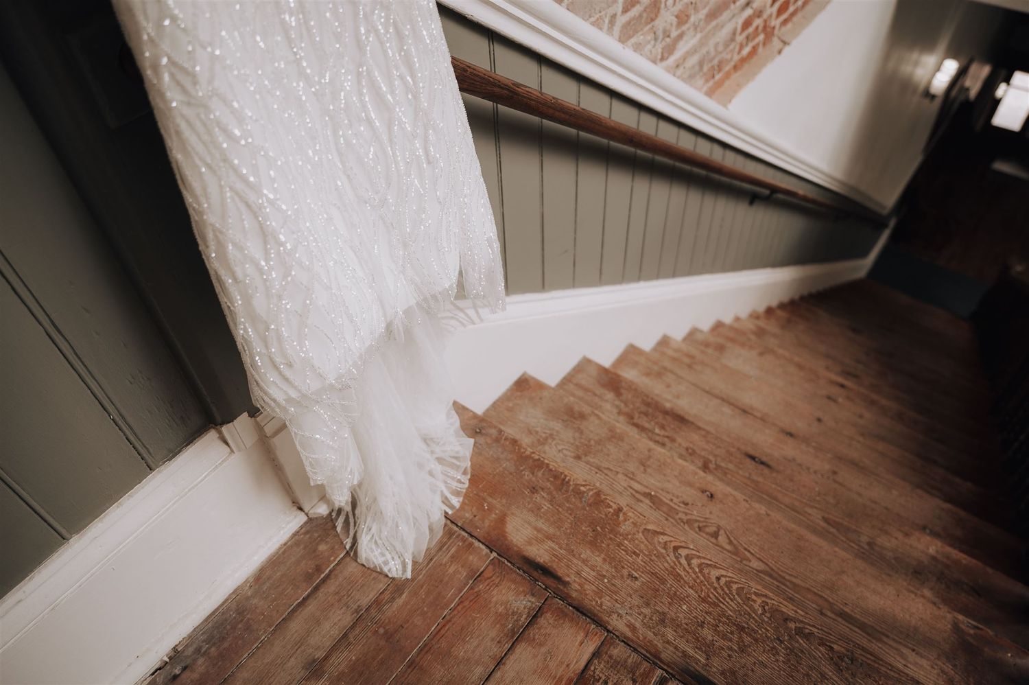 Wedding dress train flowing down wooden staircase with white baseboards and exposed brick wall.