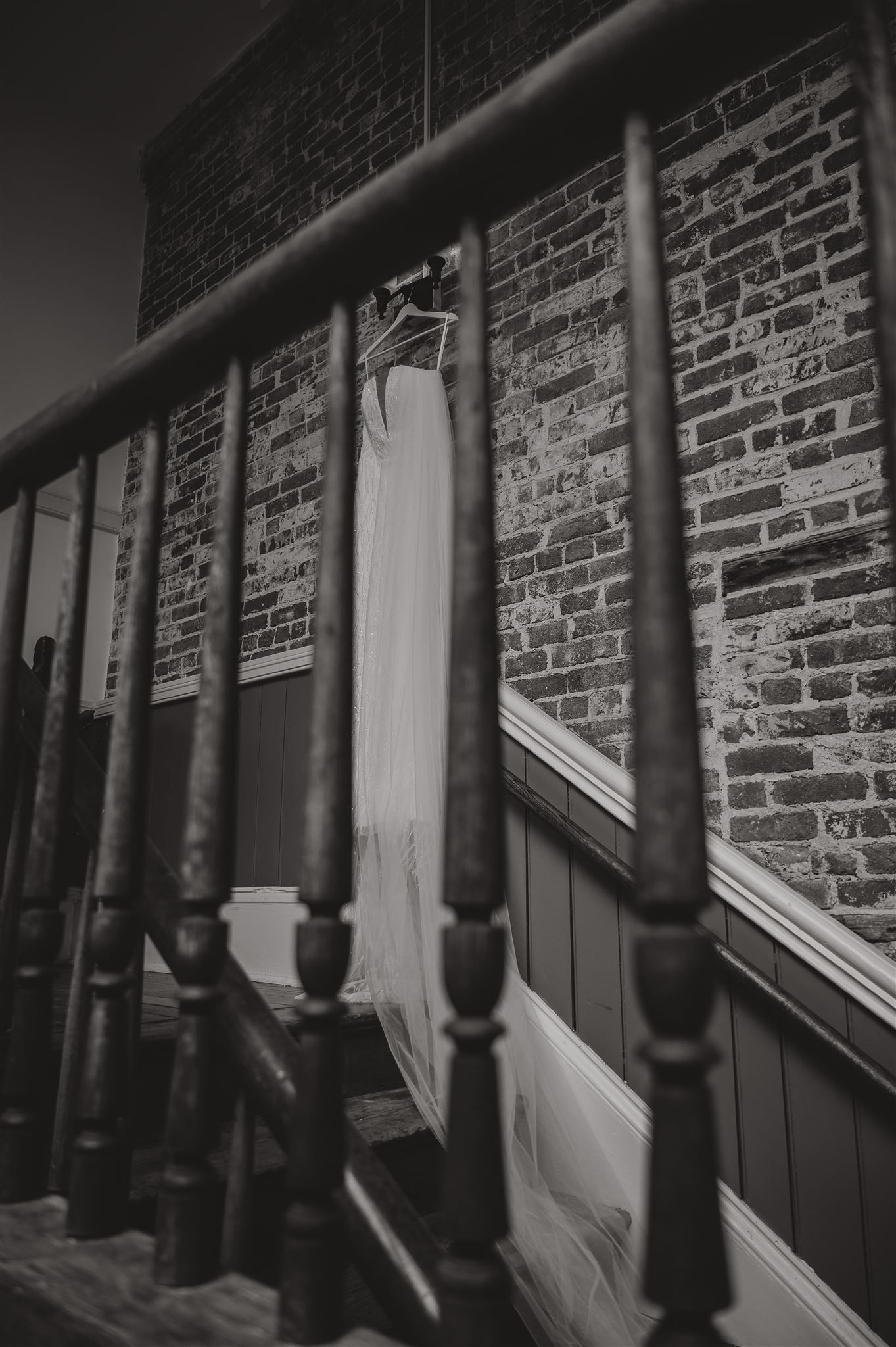 Black and white photo of wedding dress hanging by wooden staircase railing.