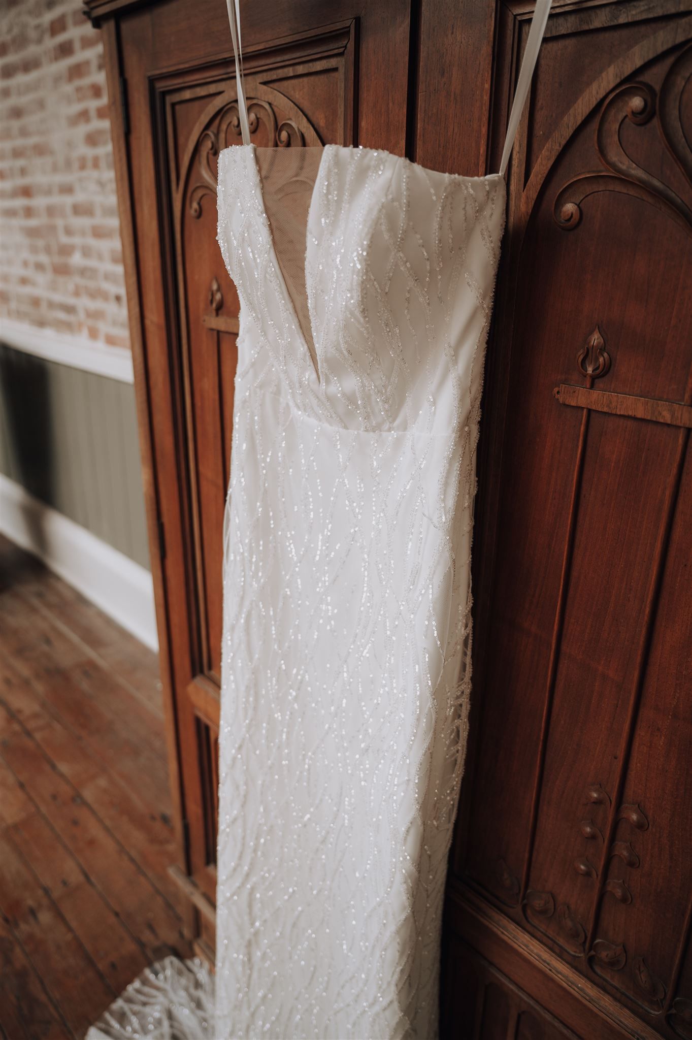 Detailed view of white wedding dress with beaded embellishments hanging against wooden door and brick wall.