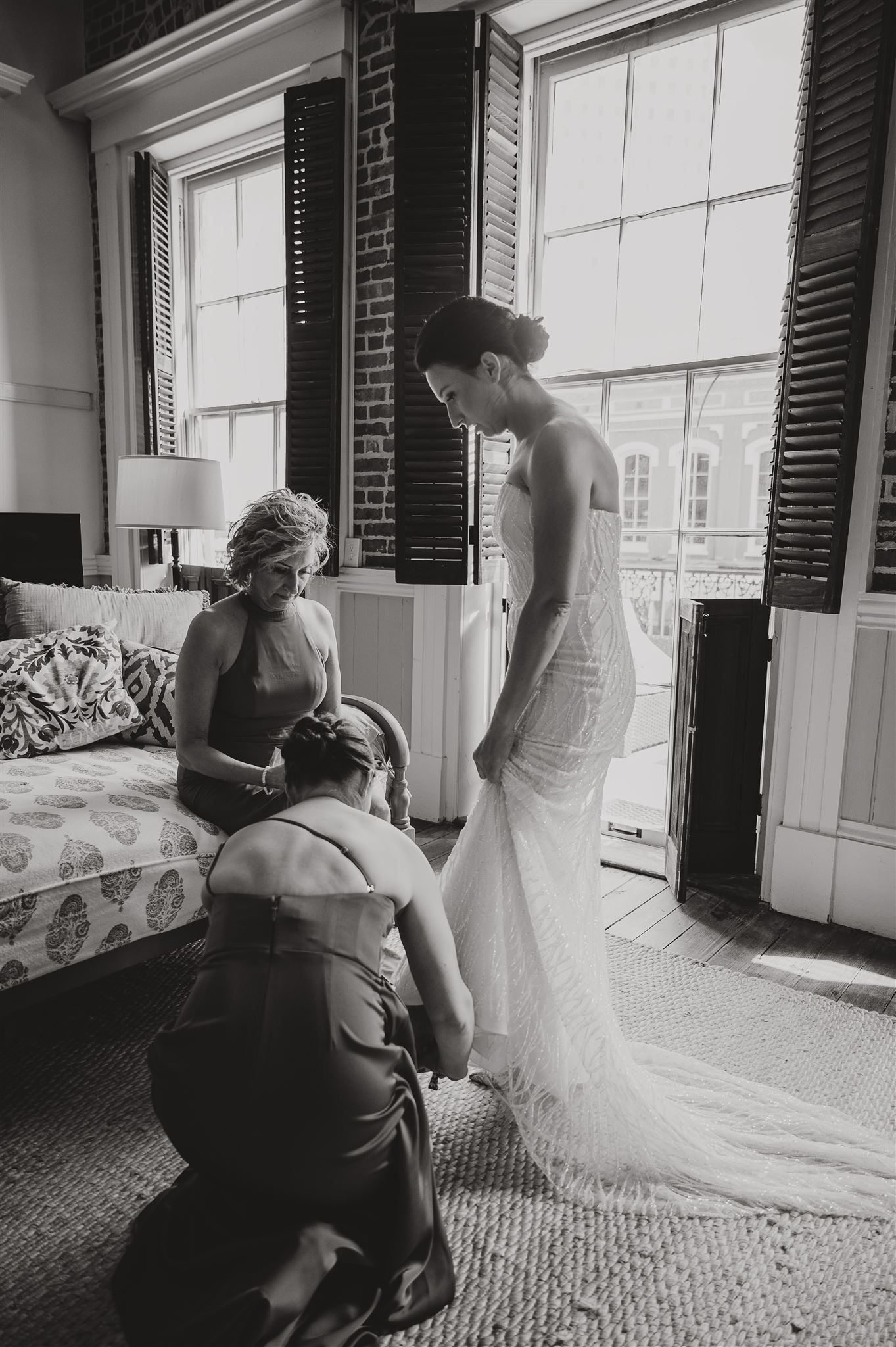 Black and white photo of wedding dress fitting in brick-walled room with natural light.