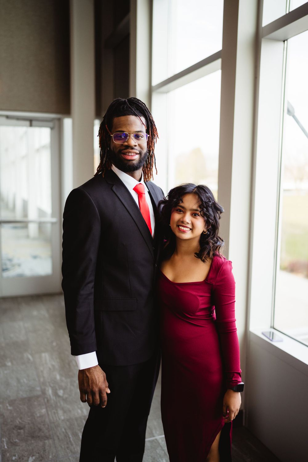 Formal couple in red and black attire posing by window.