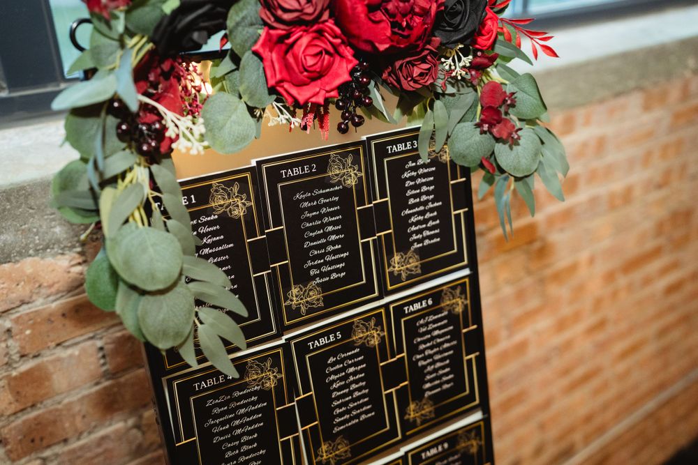 Wedding seating chart board decorated with red roses and eucalyptus against a brick wall.