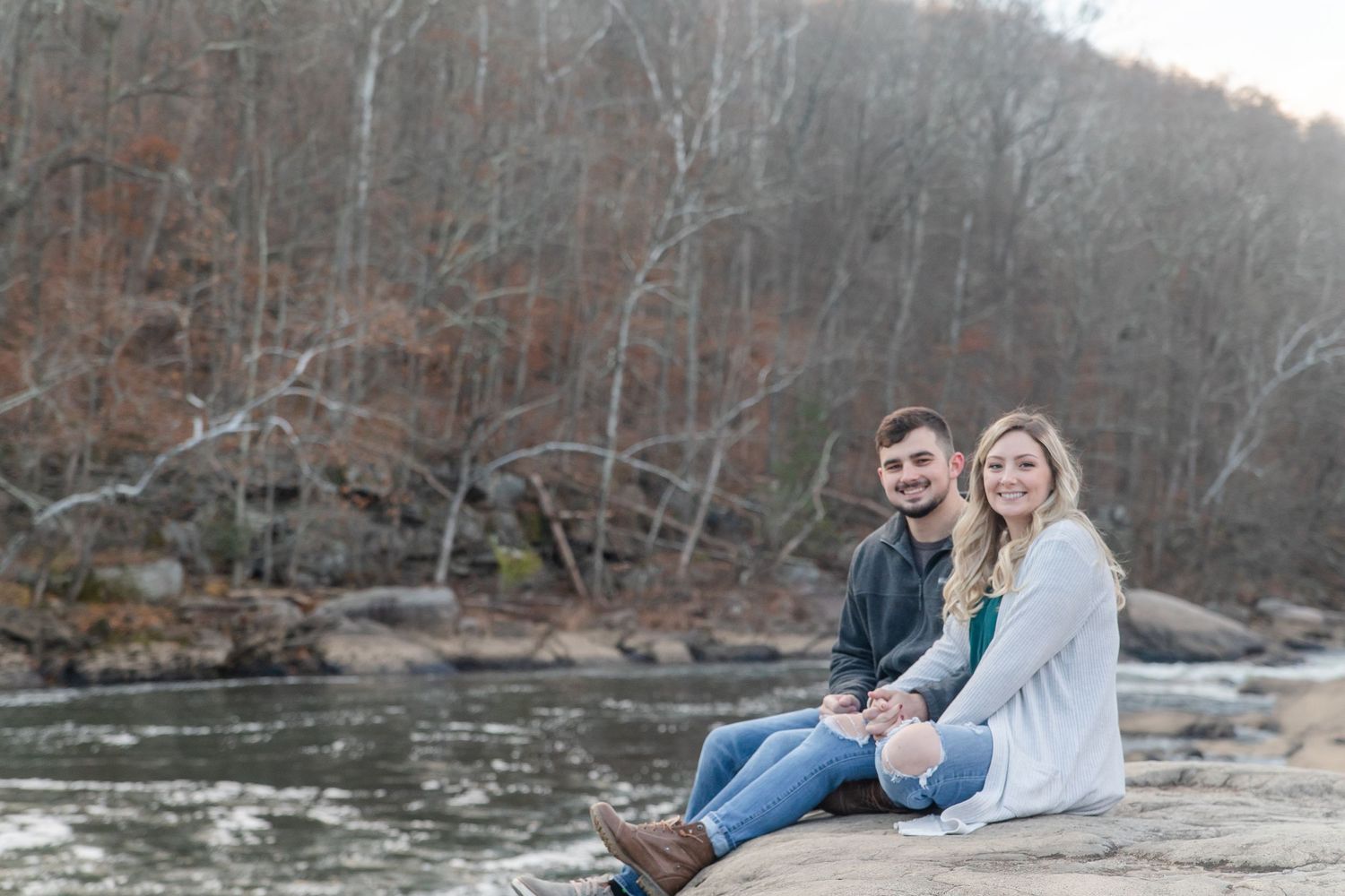 A couple relaxes by a scenic river bank surrounded by autumn foliage and rocky terrain.