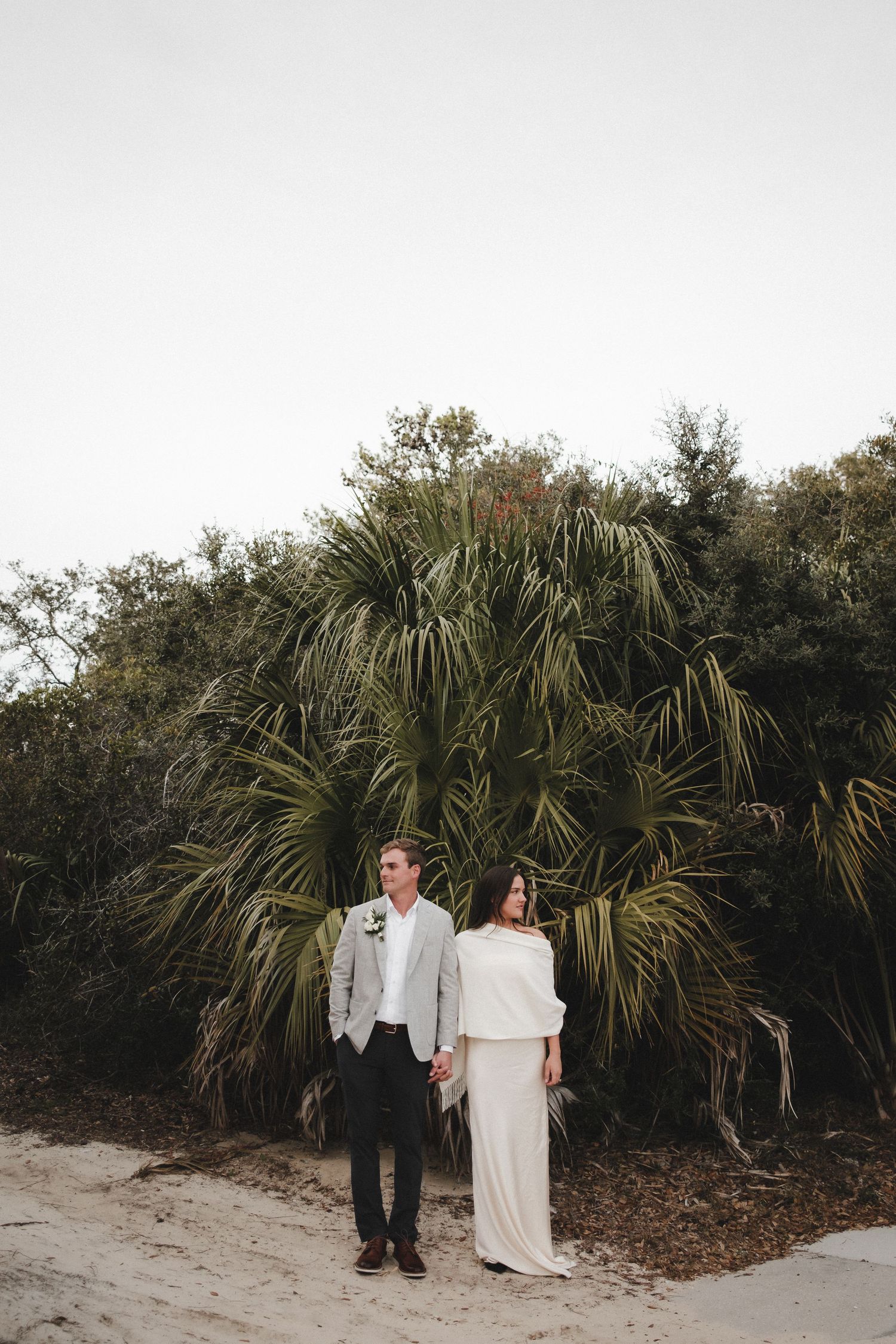 Wedding couple walking along sandy path with tropical palm trees in background at dusk.