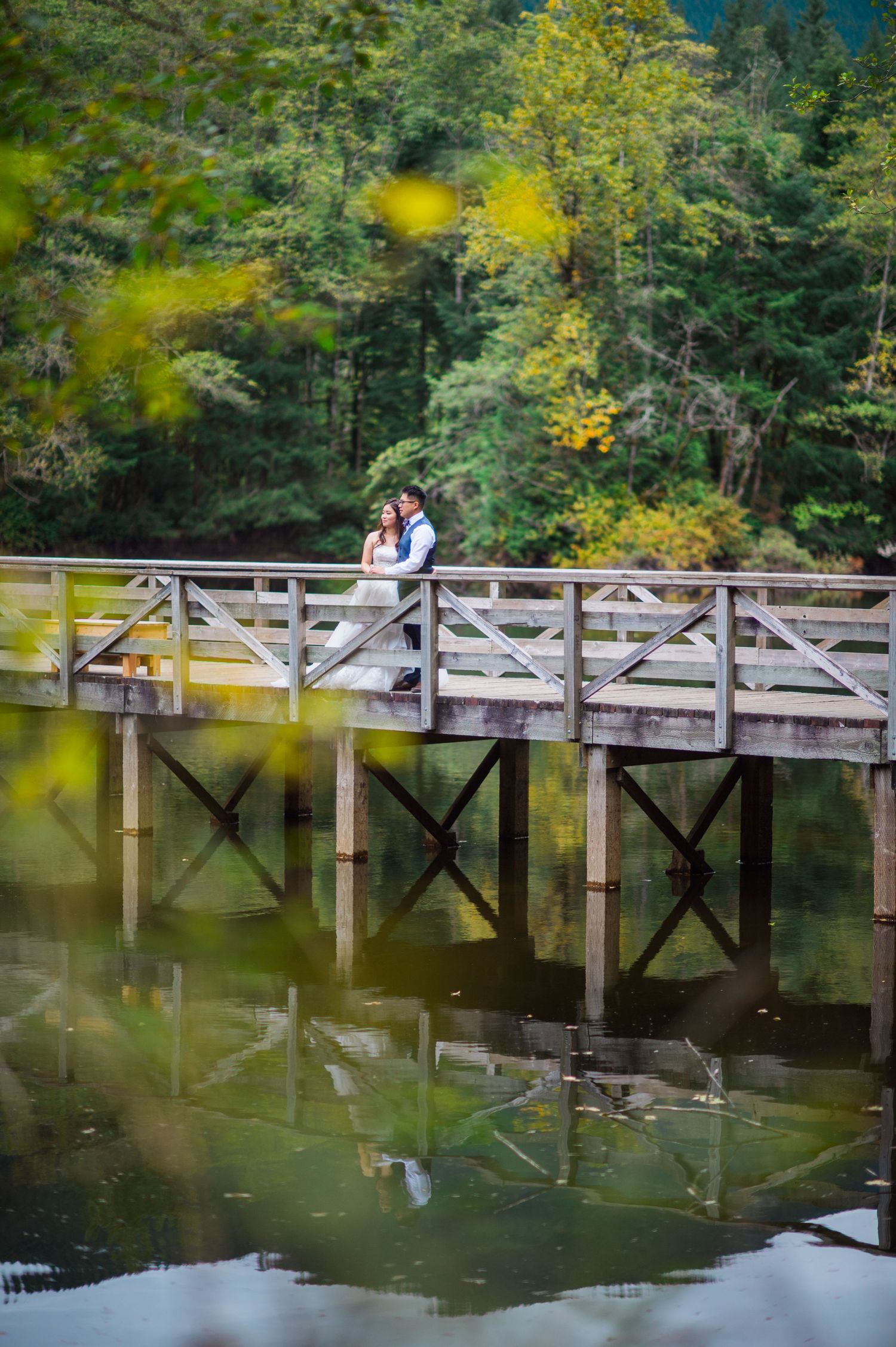 A couple stands on a wooden bridge over a calm lake surrounded by autumn foliage.