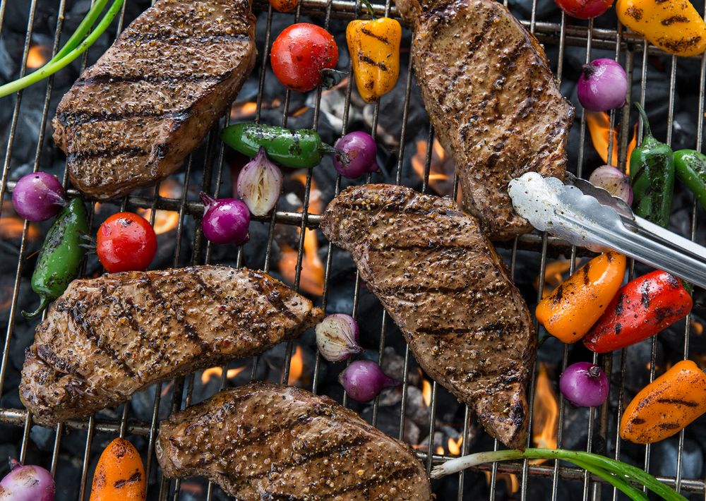 Five ribeye steaks being grilled over charcoal with and assortment of peppers, pearl onions, and cherry tomatoes.