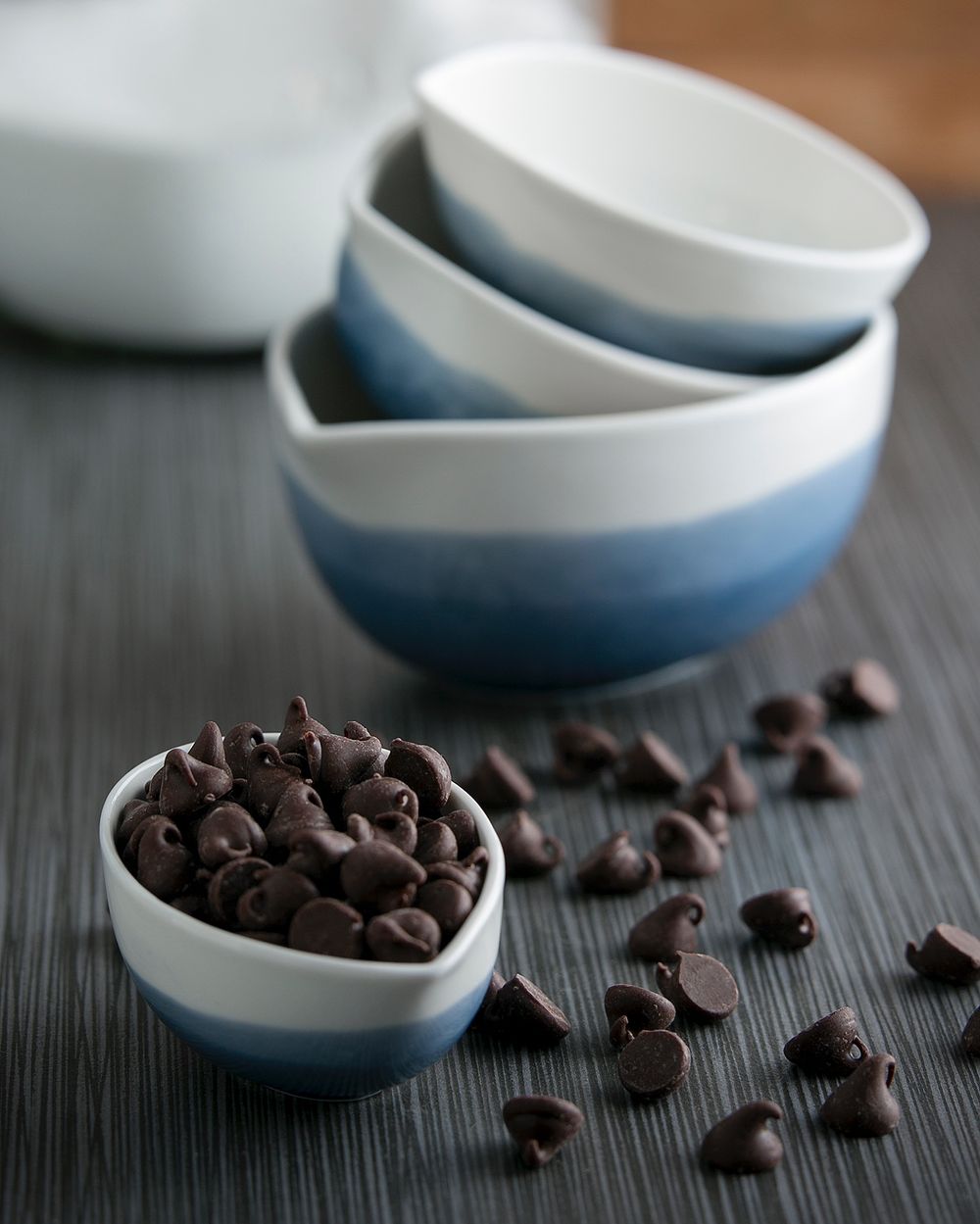 A ceramic measuring cup is overflowing with dark chocolate chips on a grey kitchen counter.