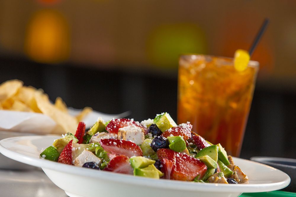 A chicken and strawberry garden salad staged with a bowl of tortilla chips and a glass of iced tea.