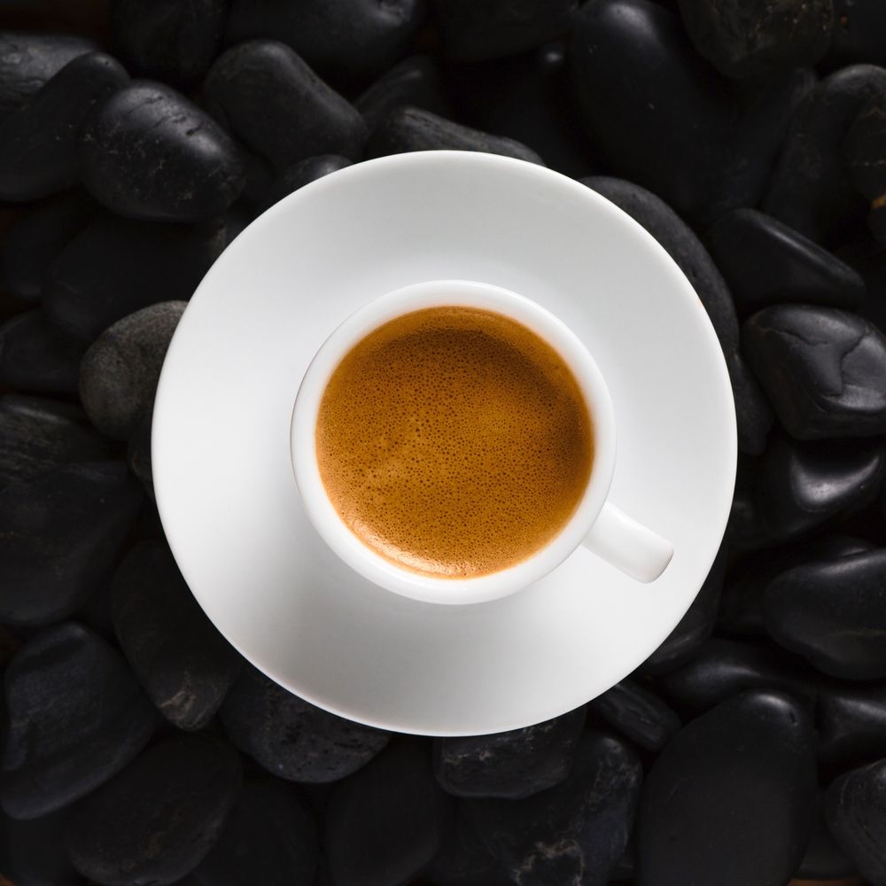 A fresh double espresso in a white demitasse mug and saucer, shot from overhead, sits on layer of black river rock.