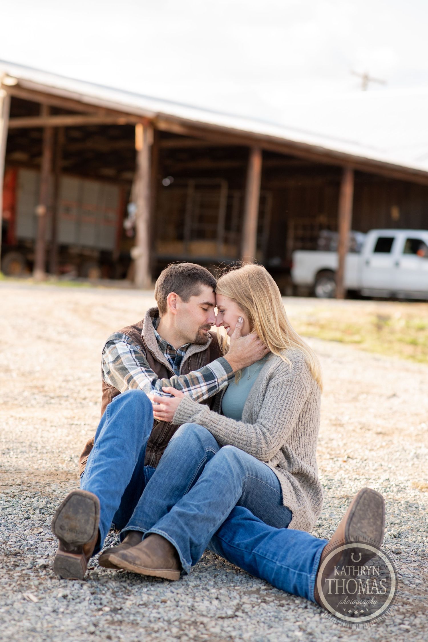 A romantic couple sitting together on gravel ground near a rustic barn building.