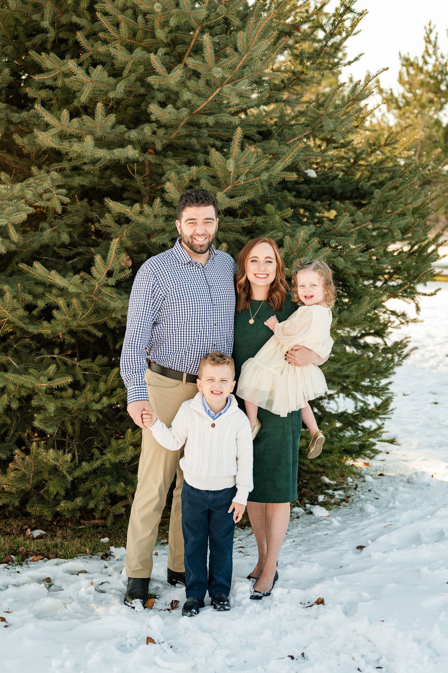 Family of four posing for a portrait in front of evergreen trees in winter snow.