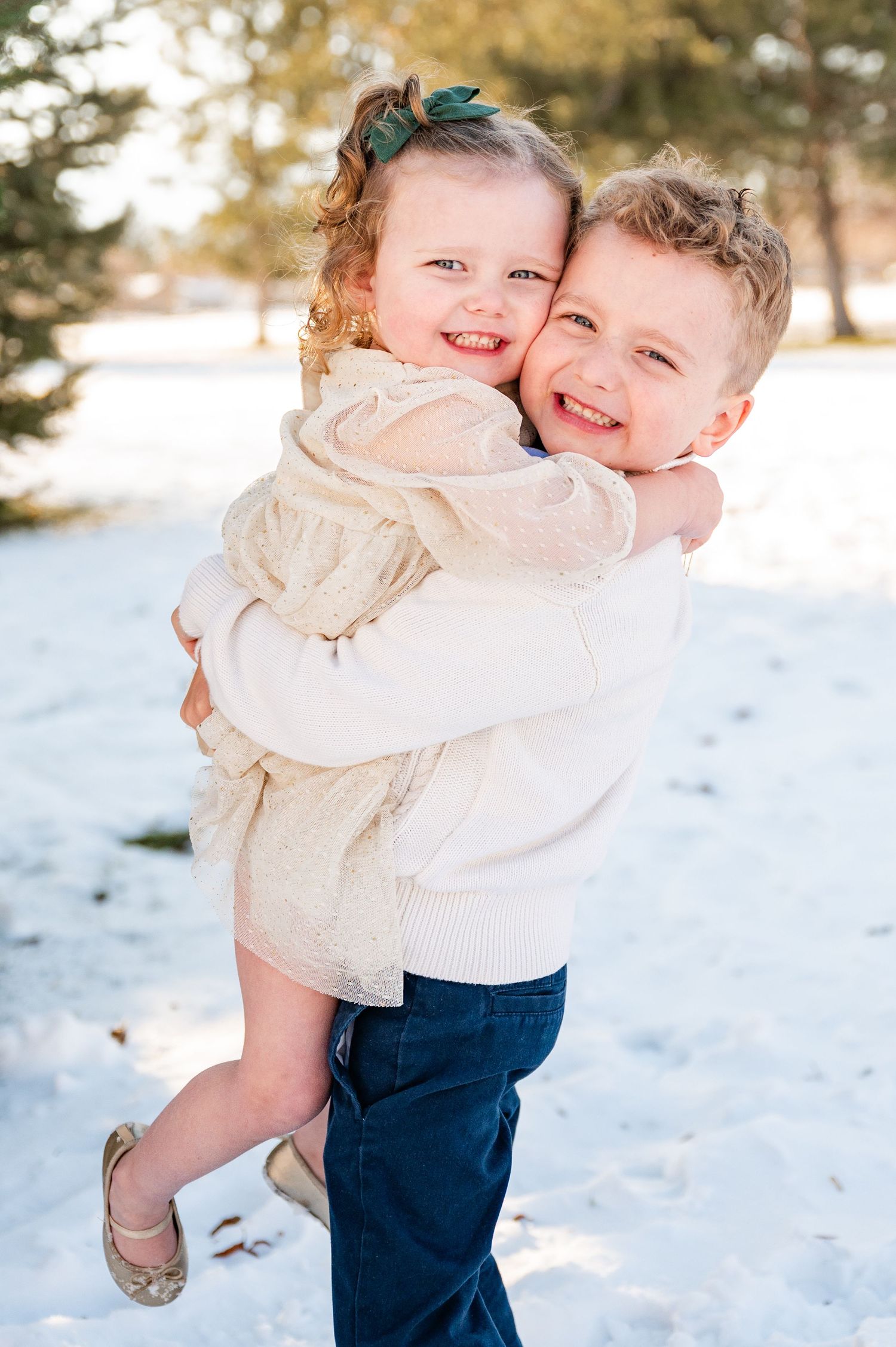 Two children hugging and smiling together in the snow wearing white sweaters.
