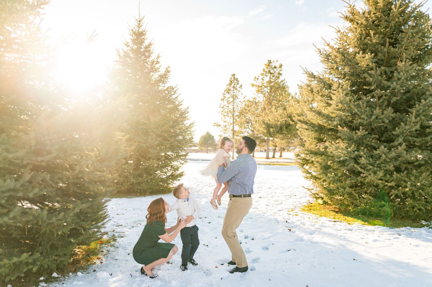 Family playing together in snowy winter scene with evergreen trees at sunset.