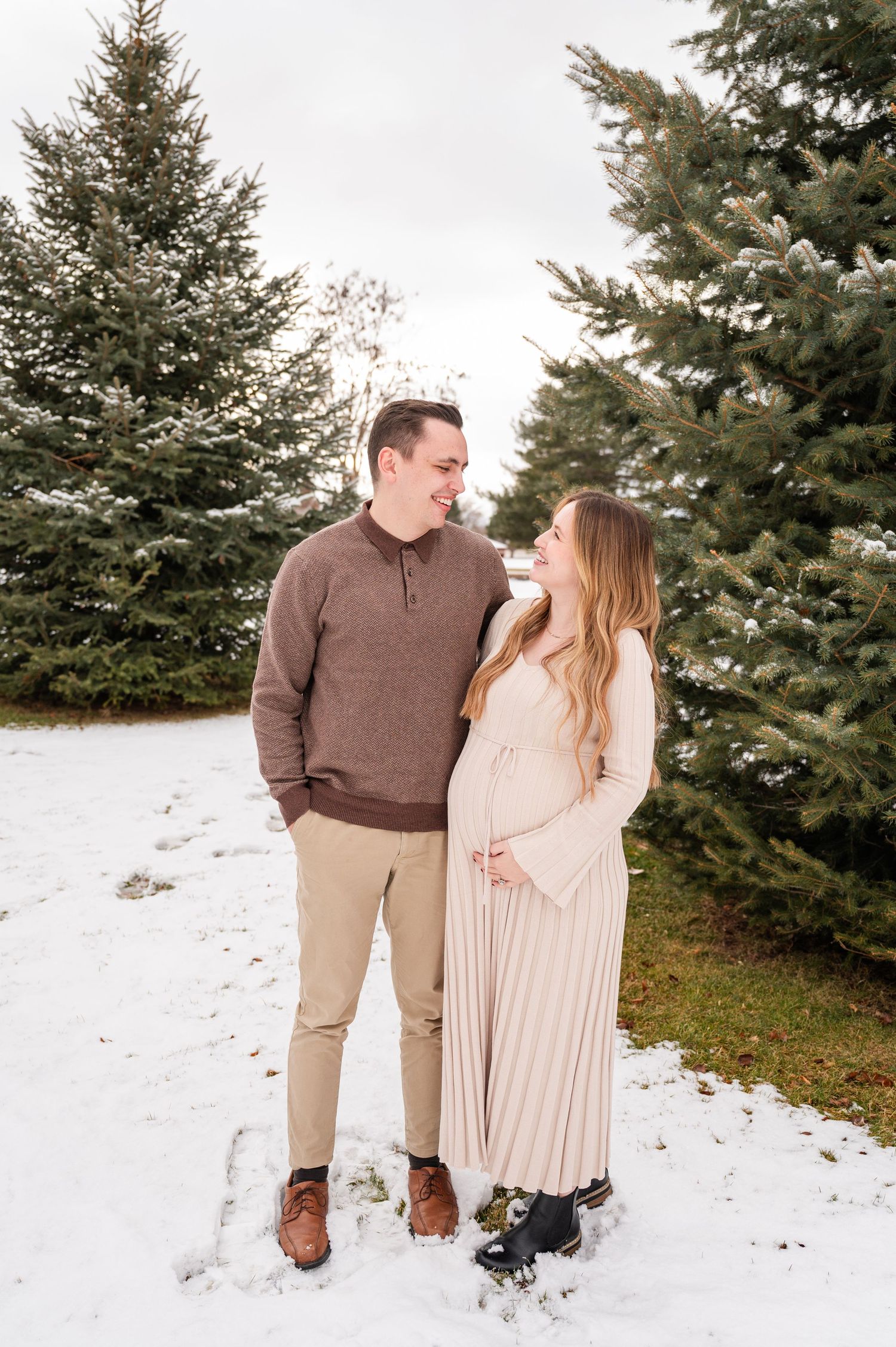 Couple stands in snowy setting among evergreen trees in winter attire.