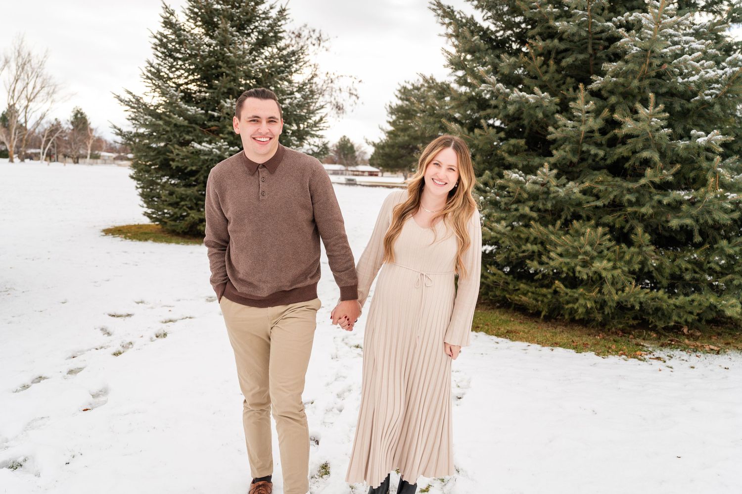 Couple holding hands while walking in snow between evergreen trees.