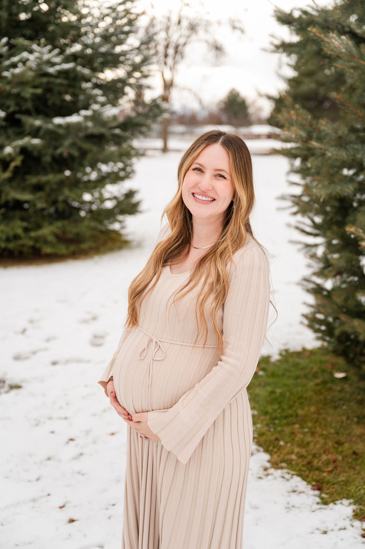 Expectant mother in beige dress smiles while standing between snow-dusted evergreen trees.