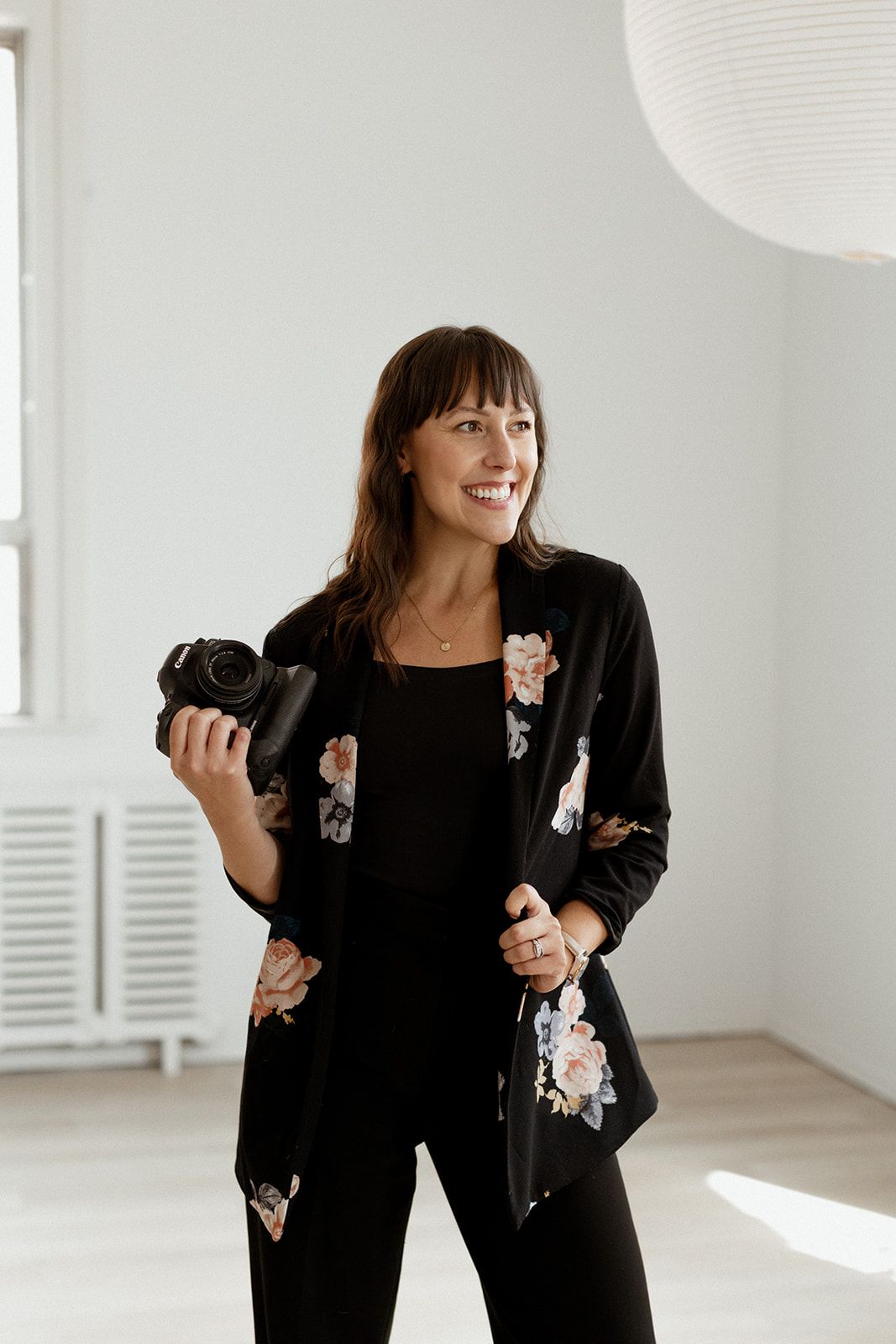 Professional photographer in floral blazer holds camera in bright minimal studio setting.