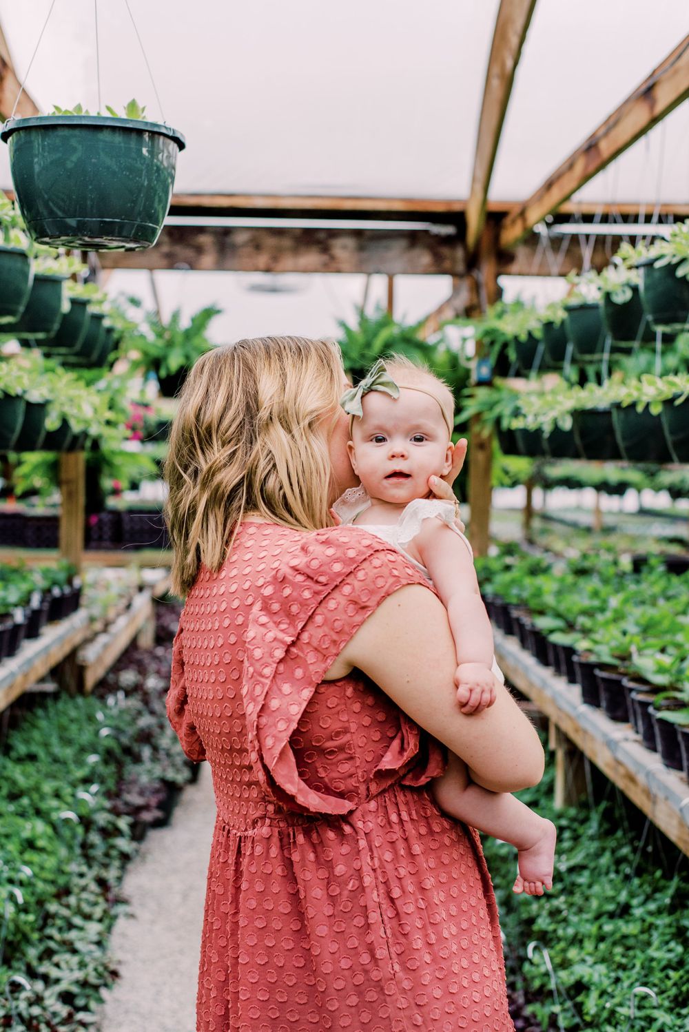 A coral dress-wearing woman holding a baby while shopping in a greenhouse nursery filled with potted plants and hanging baskets.