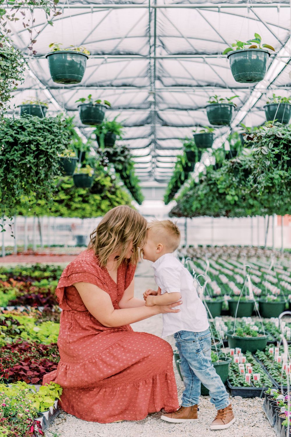 Two people share a tender moment while surrounded by potted plants and hanging baskets in a greenhouse setting.