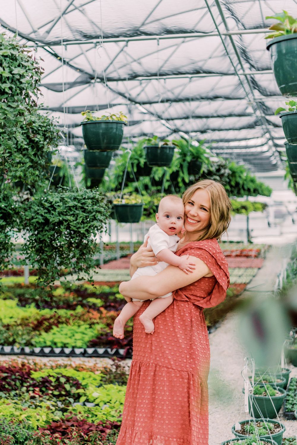Two people stand among lush plants and hanging baskets in a large greenhouse nursery environment.