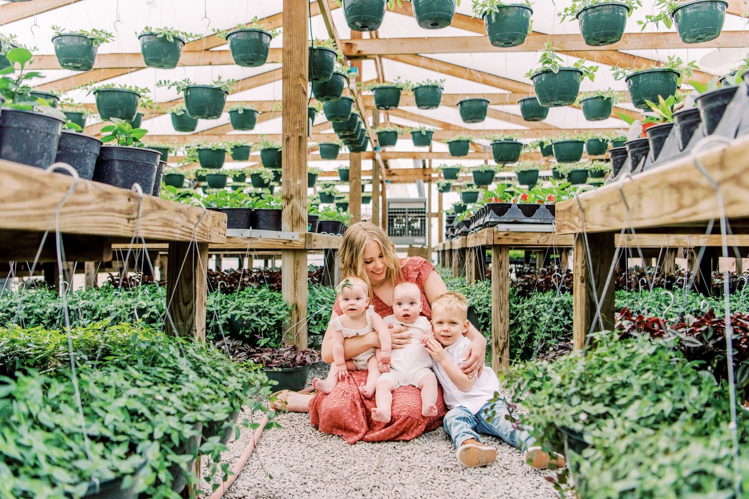 A mother sits with three young children surrounded by lush plants and hanging baskets in a bright greenhouse nursery.