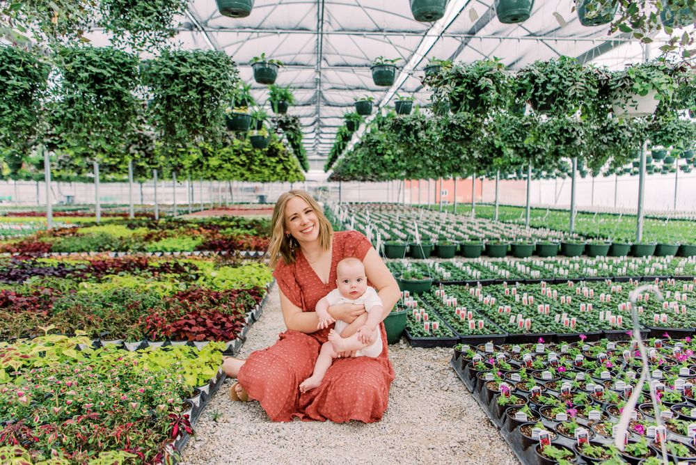 A gardener in pink attire kneels among rows of potted plants in a sunny greenhouse.