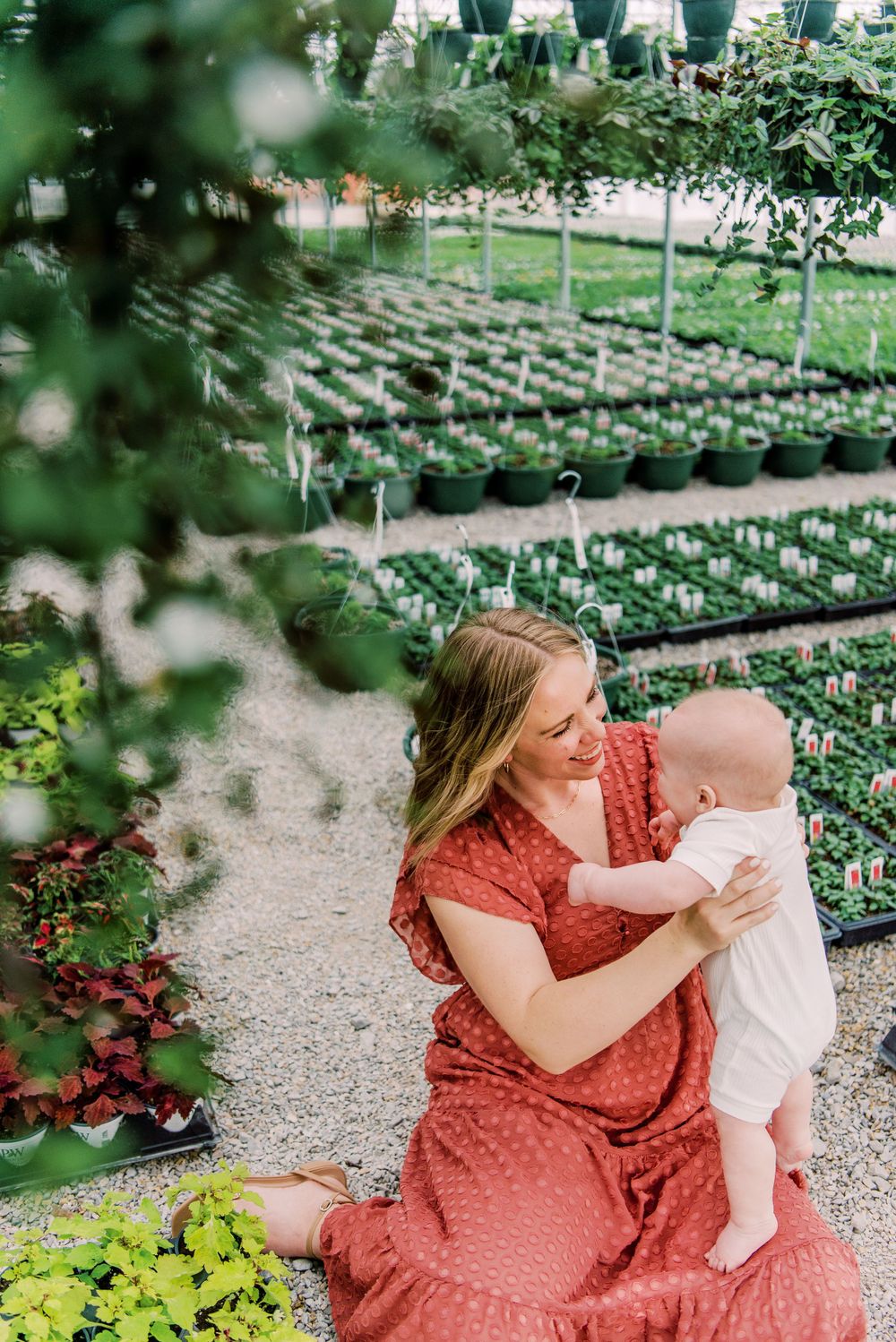 A parent in a red dress holds a baby while sitting in a garden nursery surrounded by potted plants and greenery.