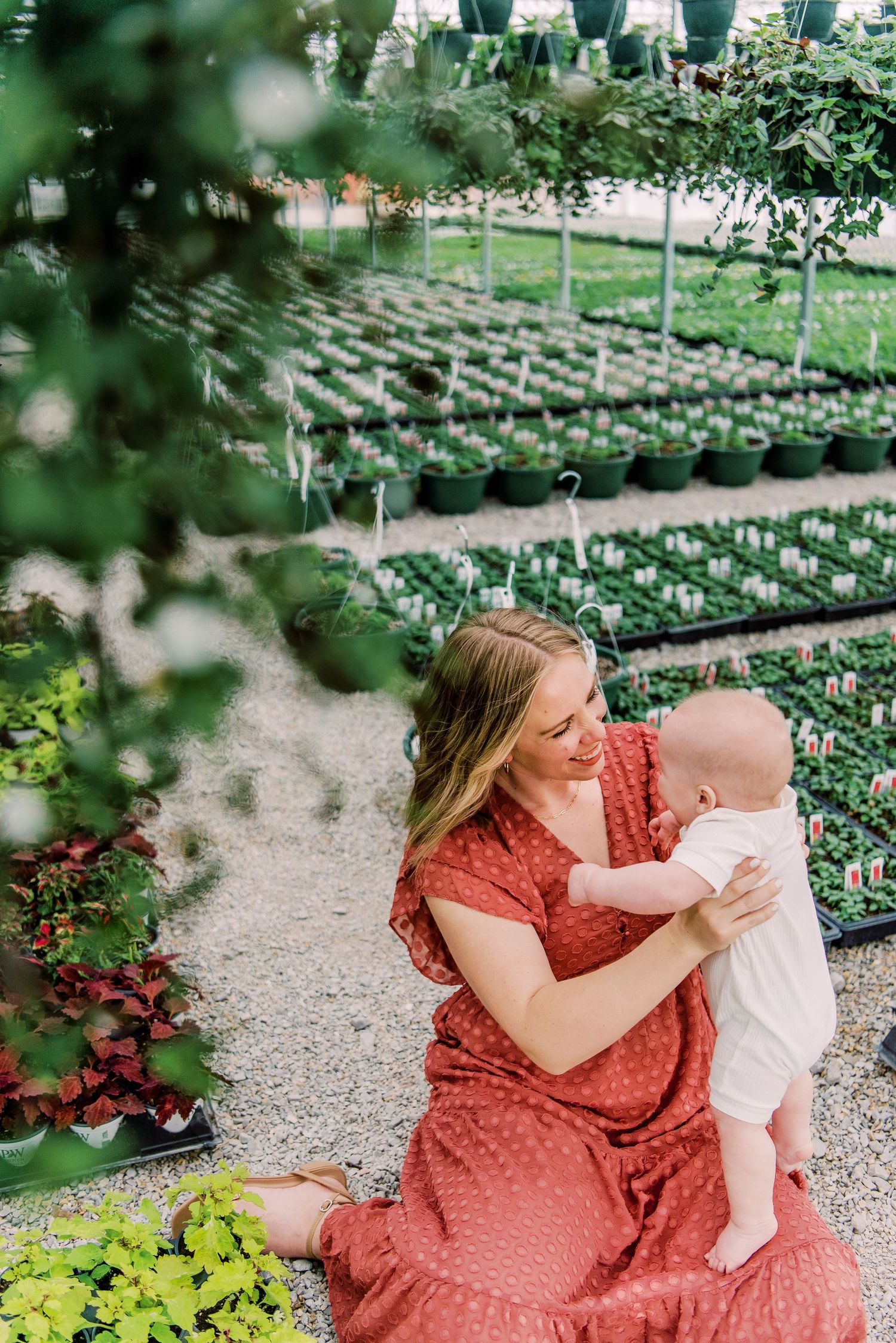 A mother in a red dress sits with her baby in a lush greenhouse surrounded by rows of potted plants.
