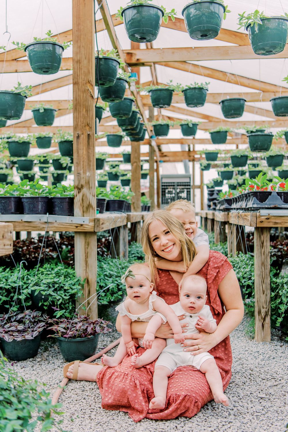 Three people sit together in a greenhouse with hanging green planters overhead and potted plants on display.