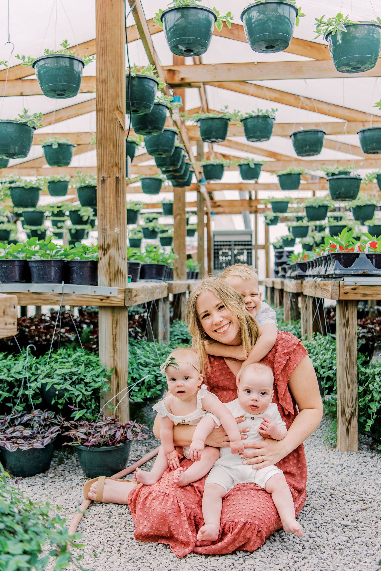 Woman in red dress sits with three babies in a lush greenhouse filled with hanging plants and greenery.