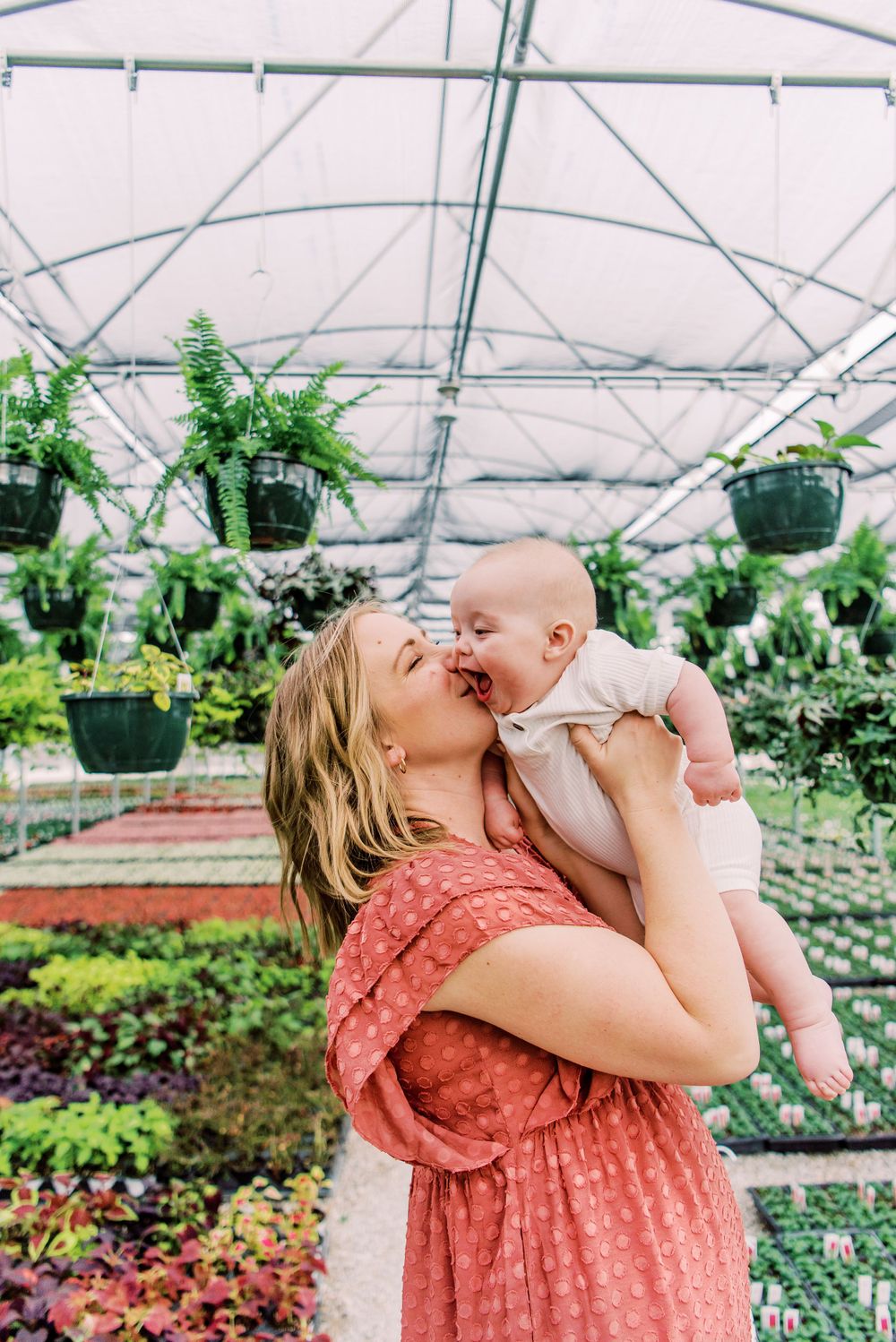 A tender moment shared between parent and baby inside a lush greenhouse filled with hanging plants and colorful flowers.