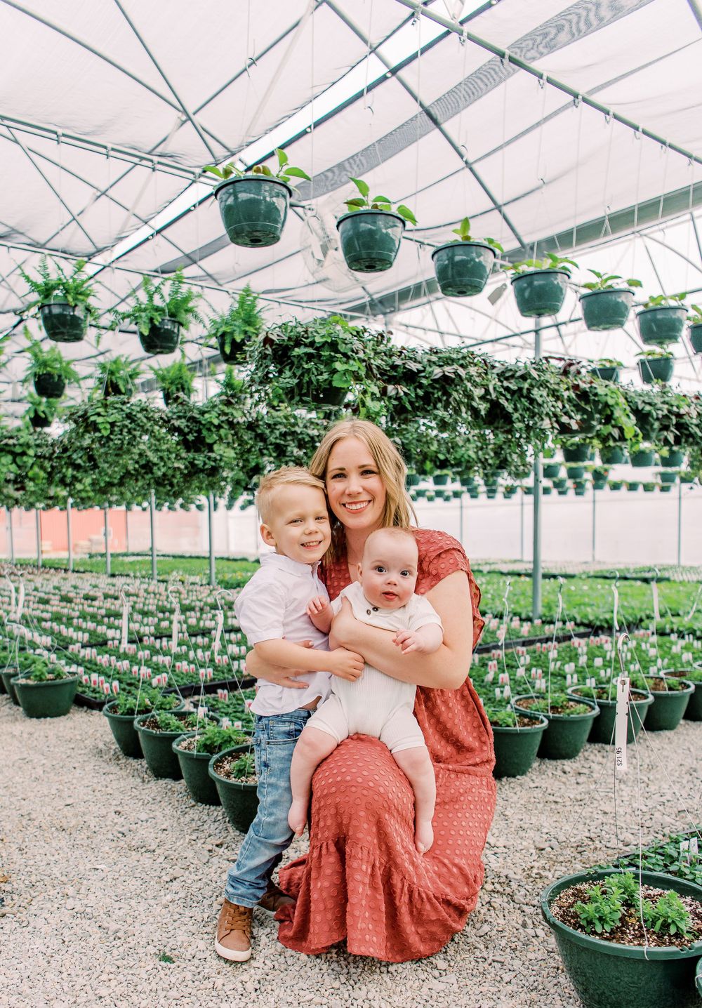 Family poses together in a greenhouse filled with potted plants and hanging baskets under industrial ceiling supports.
