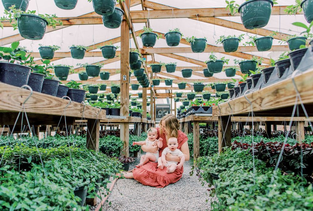 People sit among hanging planters in a greenhouse pathway lined with lush greenery under strings of teal baskets.