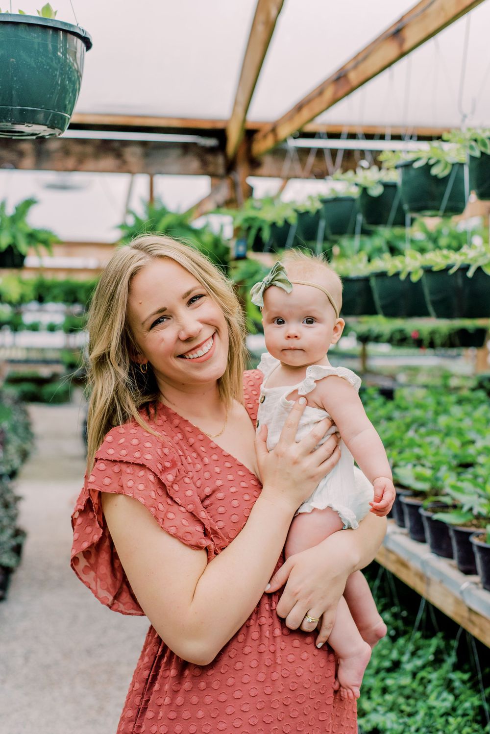 A parent holding a baby while standing in a plant nursery with green potted plants and shelving in the background.