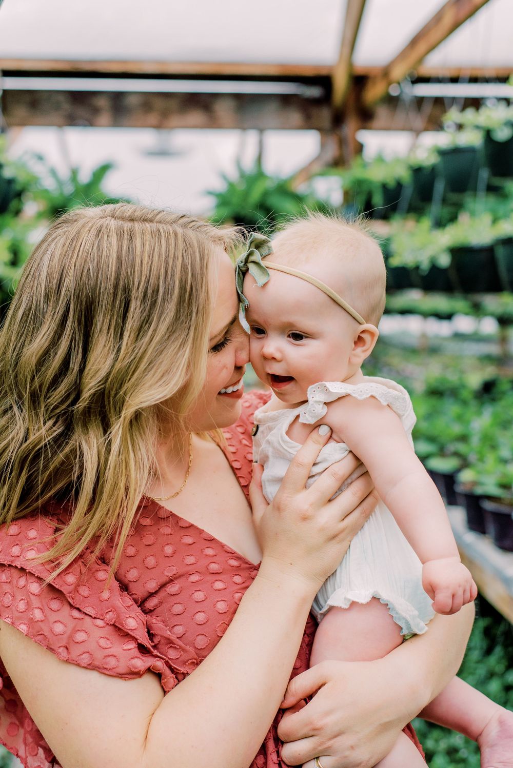 A tender moment between parent and baby in a greenhouse garden setting with natural lighting and greenery in background.